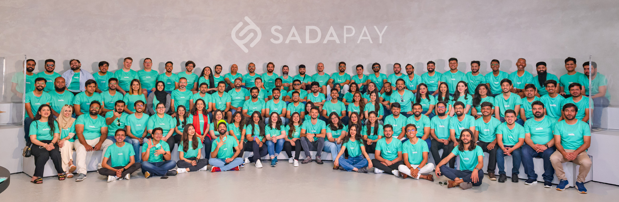 SadaPay team photo. Over a hundred employees in teal SadaPay t-shirts arranged in tiered rows under a large SADAPAY wordmark on the wall behind them.