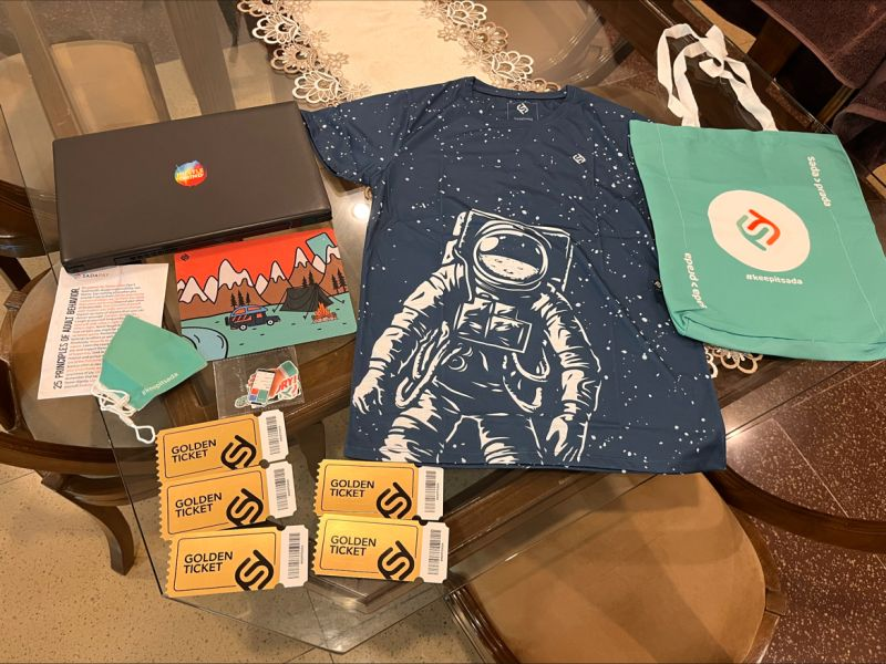 SadaPay Founders Club merchandise package: laptop sticker, navy astronaut t-shirt, teal keepitsada tote bag, postcards, behavior-principle cards, sticker pack, and five physical golden tickets
