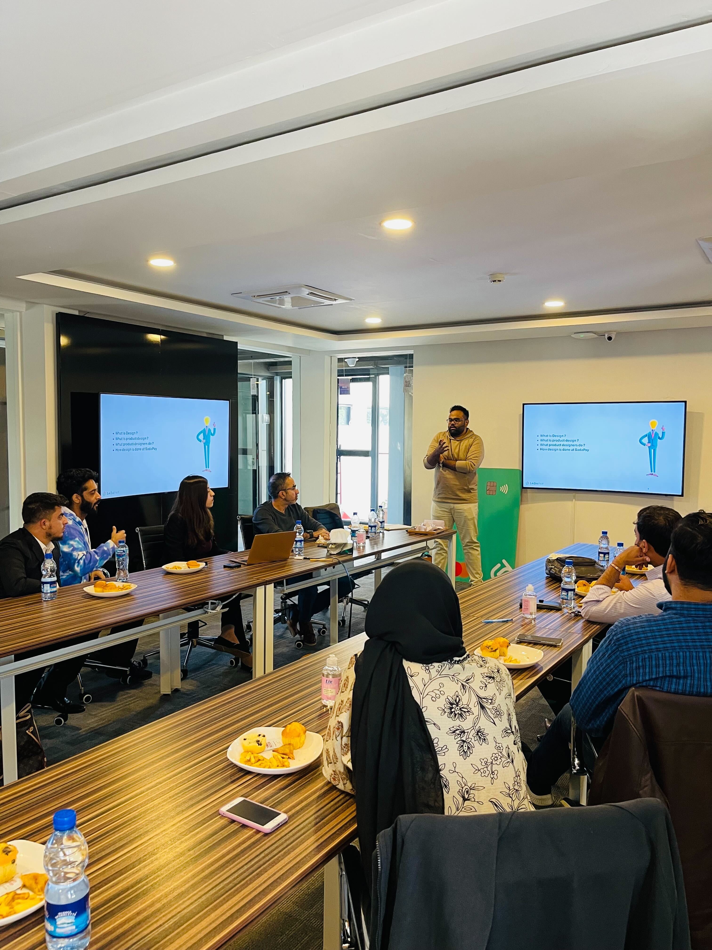 Basit Masood presenting a design culture session to SadaPay employees, with the slide deck titled 'What is Design? What is product design? What product designers do? How design is done at SadaPay.' Mixed audience around a conference table at SadaPay HQ.